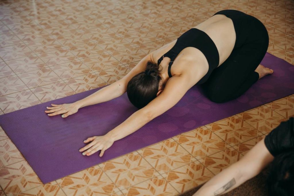 Une femme habillée en noir s'étire le dos sur un tapis de yoga mauve.