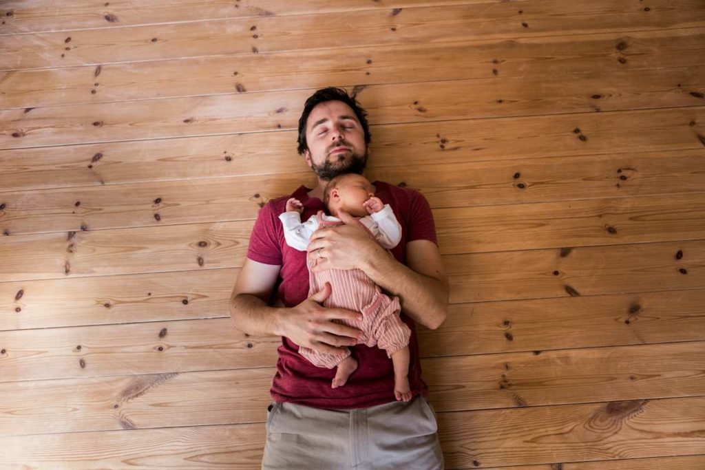 Un homme allongé sur un parquet bois en tenant son bébé dans les bras.