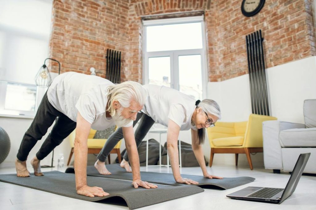 Deux personnes âgées font du yoga ensemble devant un ordinateur.