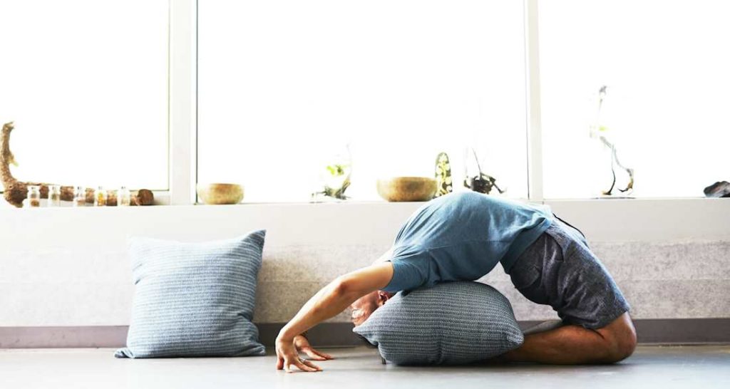 Un jeune homme effectue une pose de yoga avec un coussin dans le dos.