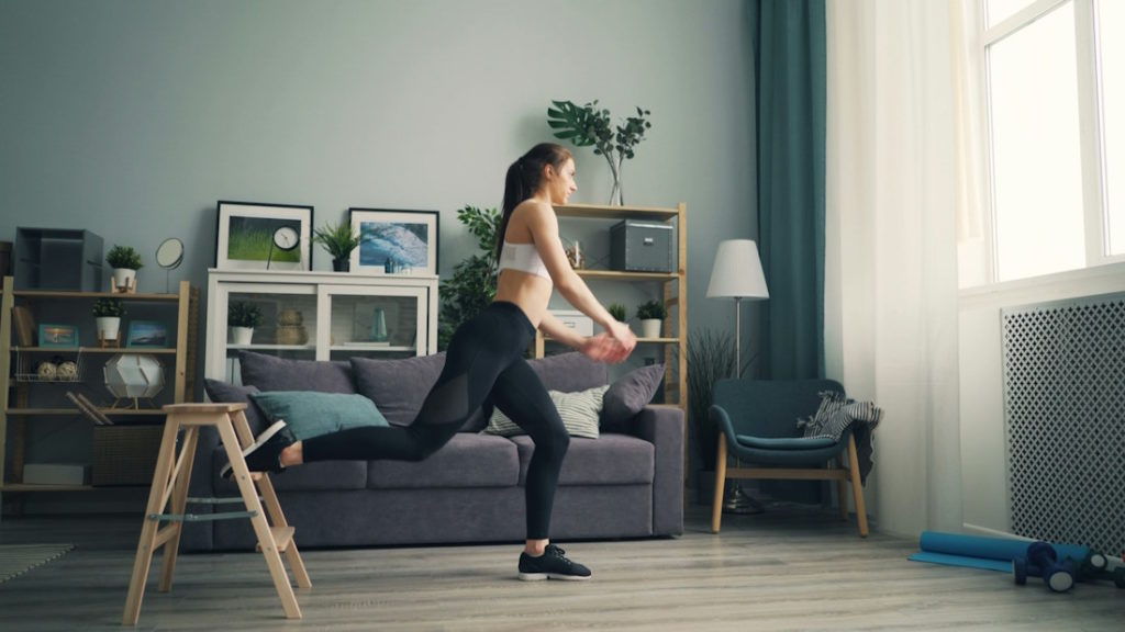 Une sportive fait un exercice de Pilates dans son salon de couleur verte, devant une fenêtre à rideaux.